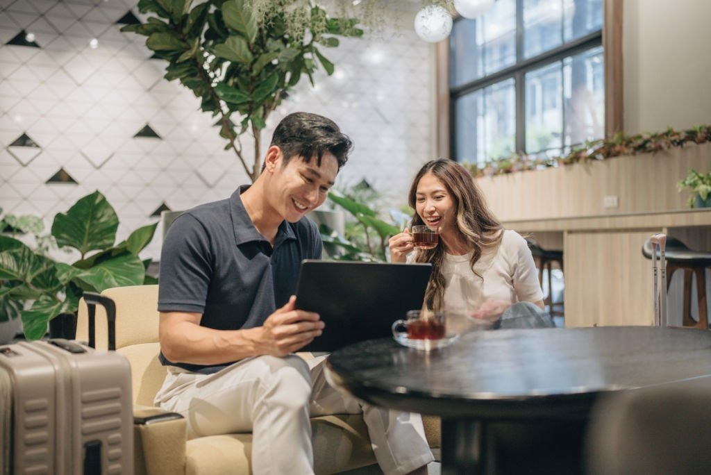 Dua karyawan memeriksa dokumen kerja di laptop sambil menunggu jadwal keberangkatan dinas