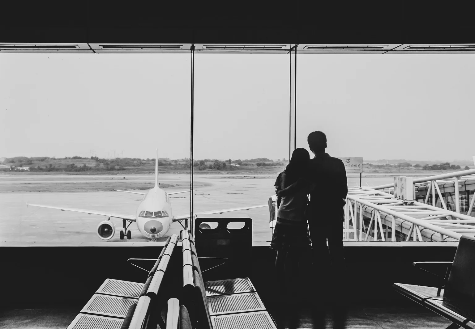 Siluet sepasang kekasih berdiri berdampingan di ruang tunggu bandara, memandang pesawat di landasan
