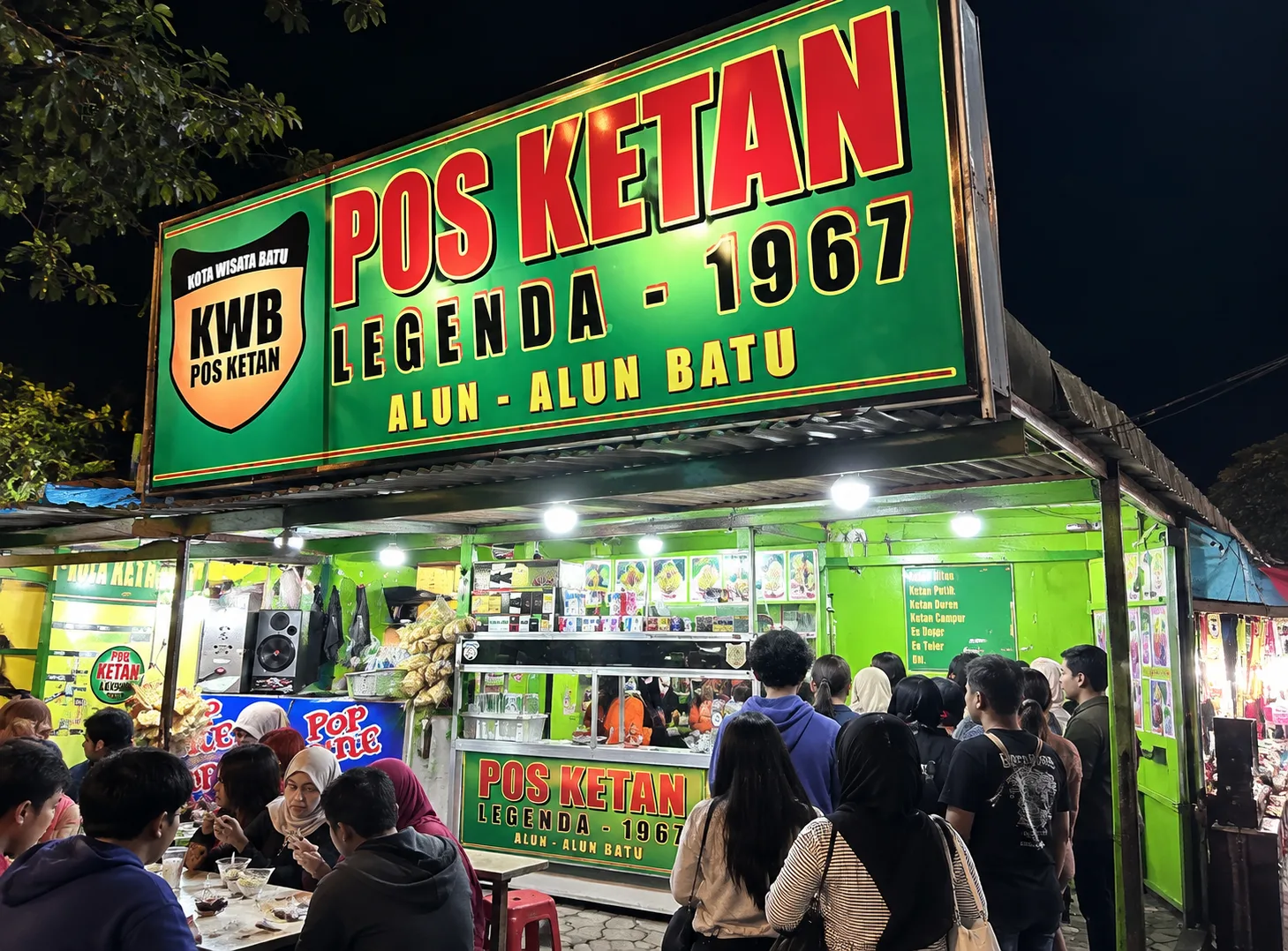 Lapak Pos Ketan Legenda 1967 di Alun-Alun Batu pada malam hari, ramai pengunjung yang antre dan makan di depan papan nama hijau