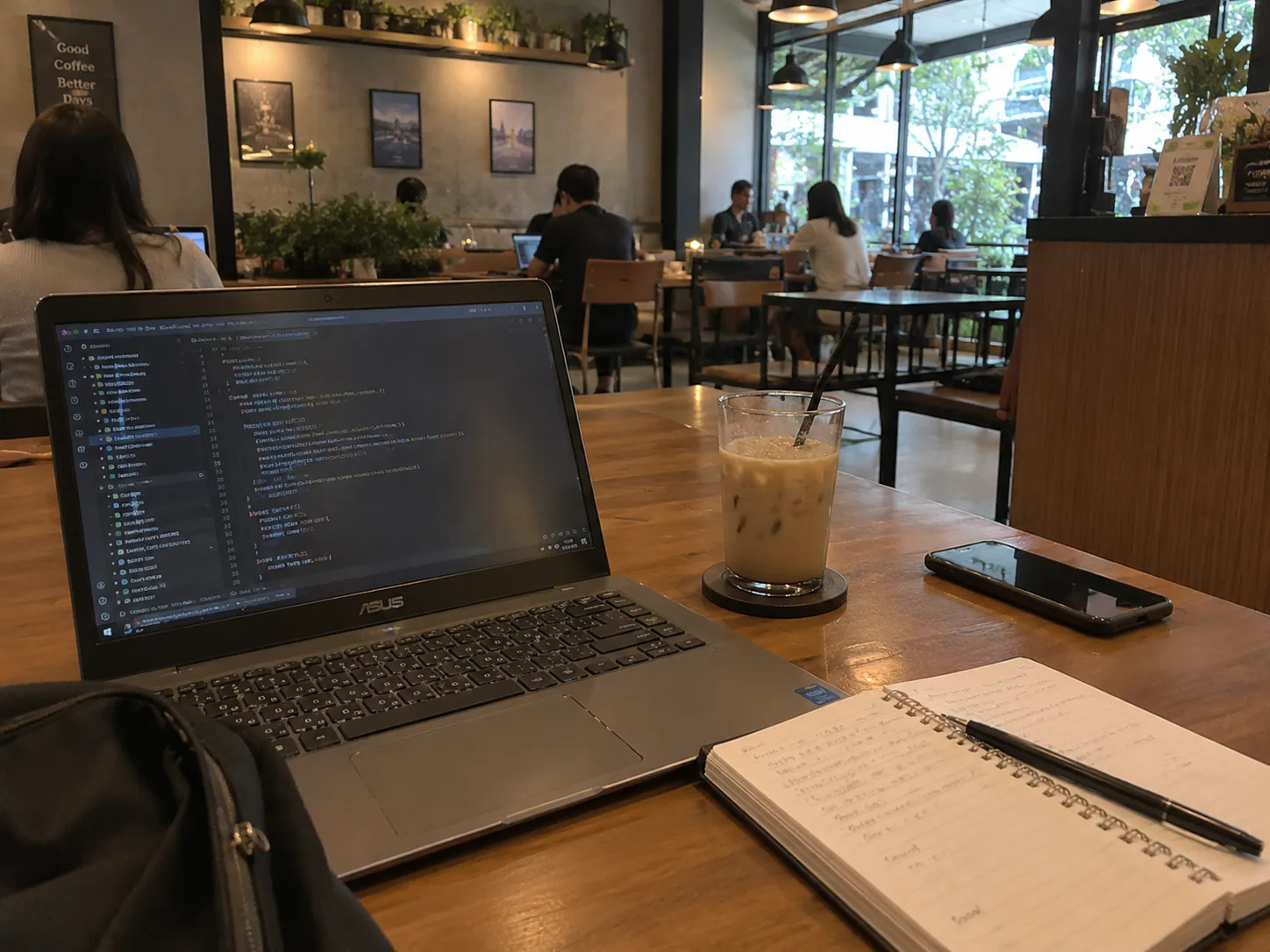 Laptop terbuka, segelas es kopi susu, ponsel, dan buku catatan di meja kayu sebuah cafe, dengan pengunjung lain yang sedang bekerja di latar belakang