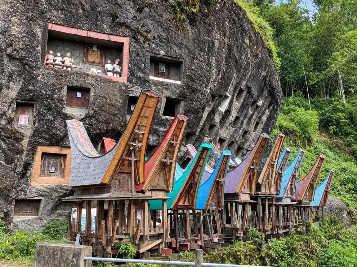 Makam pahatan dan balkon patung tau-tau di batu besar Toraja, dengan deretan bangunan beratap tongkonan warna-warni di depannya